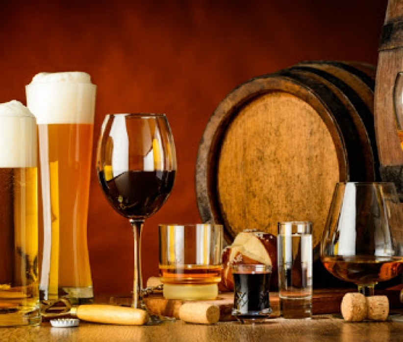 alcoholic drinks on wooden table in glasses, mugs and shots with barrel in background